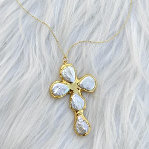 May include: A gold-toned necklace featuring a cross-shaped pendant composed of five irregular, lustrous white baroque pearls set within a polished gold metal frame. The pendant hangs from a delicate, thin gold-toned chain with a fine link design.