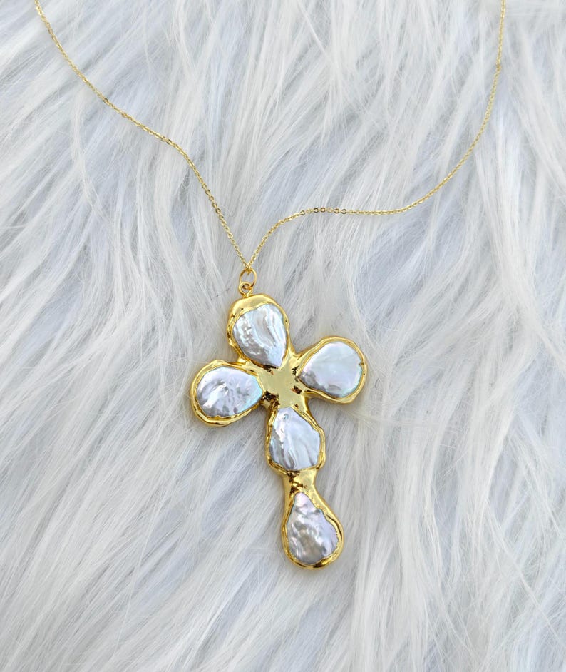 May include: A gold-toned necklace featuring a cross-shaped pendant composed of five irregular, lustrous white baroque pearls set within a polished gold metal frame. The pendant hangs from a delicate, thin gold-toned chain with a fine link design.
