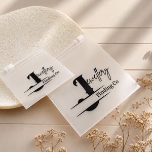 May include: Two square, semi-transparent frosted plastic pouches with white zipper closures. Each pouch features a black printed logo that reads Jewellery Finding Co with a stylized letter J. The pouches are arranged in a small and large size format.