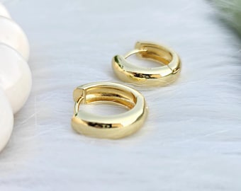 May include: A pair of small, polished gold hoop earrings with a thick, rounded profile. These earrings feature a hinged closure mechanism, showcasing a smooth, reflective metallic surface that catches the light, creating a bright and clean appearance.