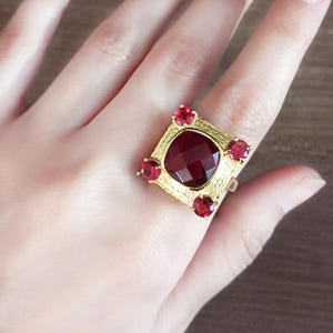 May include: A gold ring with a large, square, deep red gemstone at the centre, framed by four smaller red stones. The ring has a textured, ornate setting.