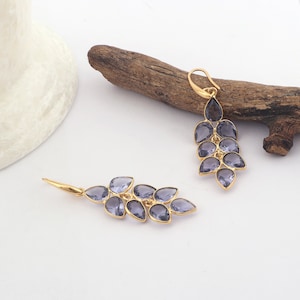 May include: Gold-toned chandelier earrings featuring a cascading design of teardrop-shaped, lavender-colored gemstones. The earrings are displayed on a white surface with a piece of driftwood in the background, showcasing the jewelry's elegant design.