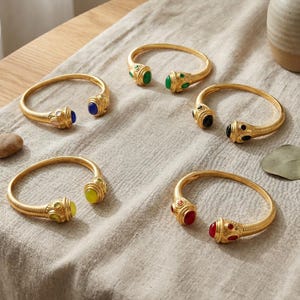 Puede incluir: Cinco brazaletes abiertos de metal dorado, cada uno con bandas texturizadas y dos terminales decorativos incrustados con piedras cabujón lisas y coloridas en azul, verde, negro, amarillo y rojo, dispuestos sobre una tela texturizada.