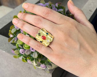 May include: A gold-toned statement ring with an organic shape, embellished with an assortment of colourful gemstones. The ring has a textured finish and is set with a red, green, blue, black, and white stone.