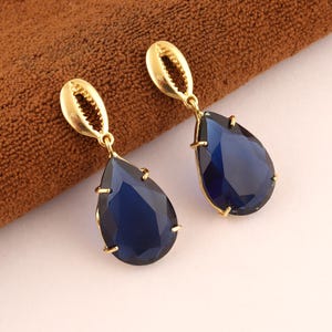Peut inclure: Une paire de boucles d'oreilles pendantes composées de pierres bleu foncé en forme de poire à facettes, serties dans des griffes en métal doré. Chaque boucle est suspendue à un élément supérieur en forme de coquillage cauri doré.