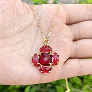 May include: A gold-colored necklace featuring a pendant with a central square-cut, deep red gemstone, surrounded by four smaller, similarly colored gemstones. The pendant is suspended from a delicate gold chain.