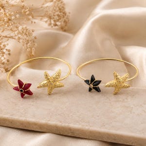 May include: Two gold-toned open cuff bracelets featuring starfish motifs. One bracelet displays a textured gold starfish and a smooth red starfish with a central black stone. The other bracelet features a textured gold starfish and a smooth black starfish.