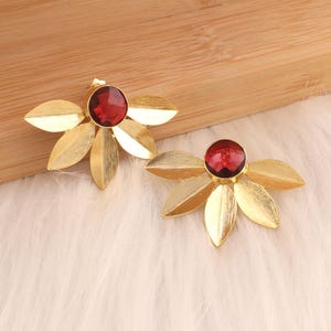 May include: A pair of gold-colored earrings with a leaf-like design. Each earring features a central, round, red gemstone. The earrings are set against a light-colored, textured background.