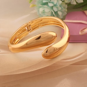 Peut inclure: Un bracelet manchette ouvert de couleur or, avec un design lisse et incurvé. Le bracelet a une charnière et une fermeture à fermoir. Le bijou est présenté sur une surface claire et texturée, avec une boîte cadeau rose et des fleurs en arrière-plan.