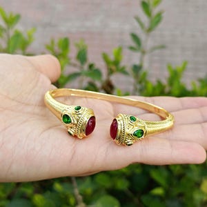 May include: A gold cuff bracelet with a snake design. The bracelet has red and green gemstones. The ends of the bracelet have a detailed, textured pattern. The bracelet is open, allowing it to be slipped onto the wrist.