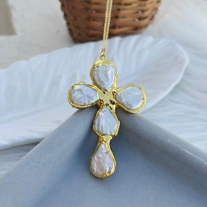 May include: A gold-toned cross pendant necklace featuring five irregular, iridescent white baroque pearls set within a textured gold metal frame. The cross is suspended from a delicate gold chain, showcasing a lustrous finish on the pearls.