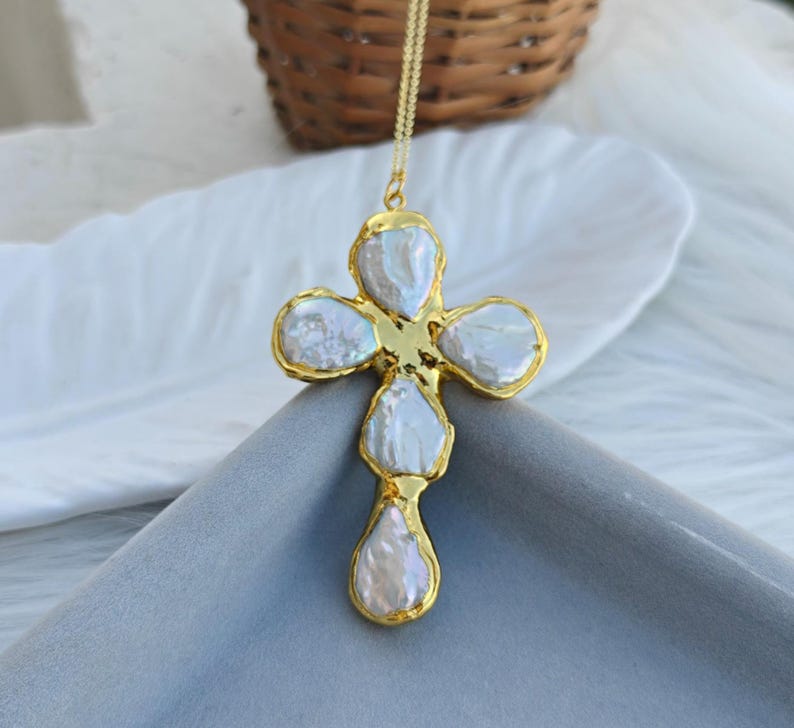 May include: A gold-toned cross pendant necklace featuring five irregular, iridescent white baroque pearls set within a textured gold metal frame. The cross is suspended from a delicate gold chain, showcasing a lustrous finish on the pearls.