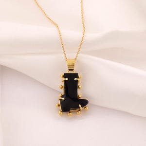 May include: A gold-coloured necklace featuring a pendant shaped like a black boot. The boot pendant is accented with small gold-coloured spheres. The necklace hangs on a gold chain against a white background.
