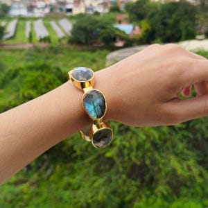 Puede incluir: Brazalete dorado con tres piedras ovaladas de labradorita iridiscente. Las piedras muestran tonos azules, verdes y grises. El brazalete se lleva en la muñeca, con un fondo verde borroso.