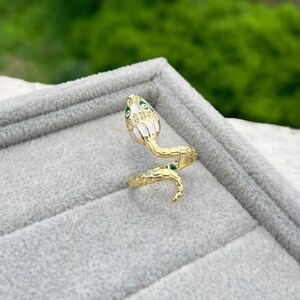 May include: A gold-coloured snake ring with a white and green enamel design. The snake's head features green eyes and a white and gold pattern. The ring is set against a grey surface with a blurred green background.