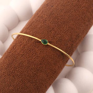 May include: A thin gold-toned bangle bracelet featuring a single, small, teardrop-shaped green gemstone centered on the band. The metal has a smooth, polished finish, and the green stone is set in a simple, minimalist metal bezel setting.