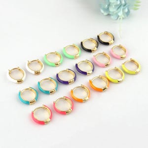 Puede incluir: Una colección de pendientes de aro dorados con detalles de esmalte de colores. Los pendientes presentan una variedad de colores brillantes, incluyendo blanco, verde, negro, rosa, amarillo, morado, turquesa, naranja y rojo. Los pendientes están dispuestos sobre una superficie blanca.