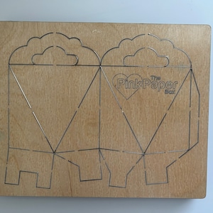 May include: A wooden die-cut template for creating a paper box. The template features a heart with the text "The Pink Paper Box" and a cloud design.