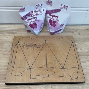 May include: Two white heart-shaped boxes with pink and purple designs. The boxes have the text "Happy Valentines Day" and "To Kennedy From Jameson". A wooden die-cut template for making the boxes is also shown. The template has the text "The Pink Paper Box".