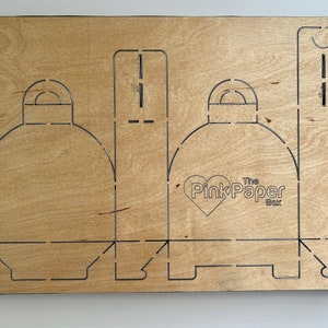 May include: A wooden template for a box with a heart-shaped logo that says "The Pink Paper Box". The template has cutouts for folding and assembling the box.