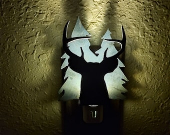 May include: A metal deer silhouette nightlight with a light bulb inside. The deer is surrounded by two pine trees. The nightlight is plugged into an outlet.