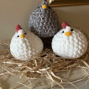 May include: Three crocheted chickens in a nest of straw. One chicken is gray and white, and the other two are white with pink and yellow accents.