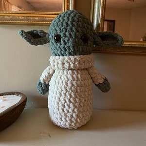 Op de afbeelding: Een gehaakt pluche speelgoed dat lijkt op een personage uit de Star Wars-franchise. Het speelgoed is groen met grote ogen en witte vacht op het lichaam en de armen.