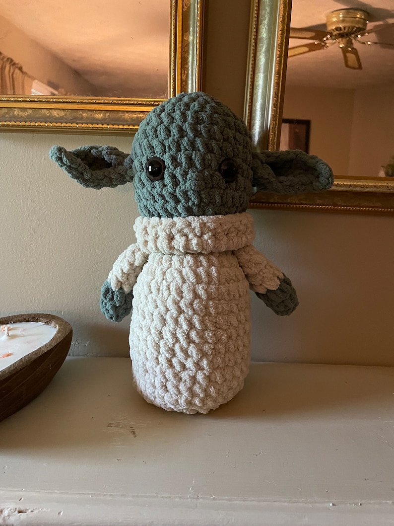 Op de afbeelding: Een gehaakt pluche speelgoed dat lijkt op een personage uit de Star Wars-franchise. Het speelgoed is groen met grote ogen en witte vacht op het lichaam en de armen.