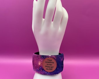 Pulsera AAC «Tengo algo que decir» para iniciar la comunicación, funda de tela sobre lazo elástico negro brillante y etiqueta de polipiel rosa.