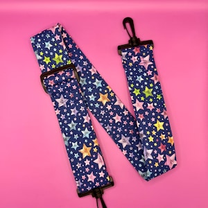 May include: A navy blue bag strap featuring a pattern of colourful stars. The strap has black plastic clips and a buckle for adjustment. The stars are in shades of pink, yellow, green, and blue.