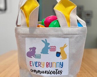 Cesta de Pascua con orejas de conejo amarillas para difundir un mensaje inclusivo estas fiestas, dispositivos de CAA y libro.
