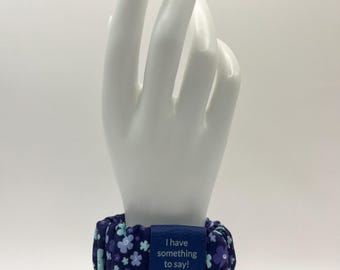 Pulsera ancha de flores estilo coletero AAC «Tengo algo que decir» para iniciar la comunicación, con etiqueta de polipiel grabada de color azul a plateado.