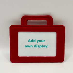 May include: A red, briefcase-shaped display frame with a handle at the top. The frame has a white interior with the text "Add your own display!" in teal. The frame is designed to hold a small display.