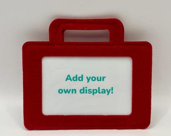 Dispositivo de comunicación aumentativa y alternativa (CAA) de fieltro con ventana para escribir mensajes y celdas insertadas del tamaño de un elfo navideño u otros muñecos. Representa la CAA en exhibiciones o juegos de rol.