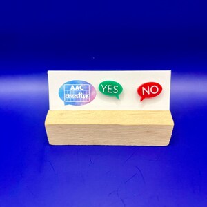 May include: A wooden block with a white sign featuring the text "AAC creative" in a speech bubble, a green speech bubble with "YES", and a red speech bubble with "NO". The background is a solid blue.