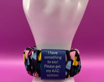 Pulsera ancha estilo coletero AAC «Tengo algo que decir» para iniciar la comunicación, con etiqueta de polipiel grabada de color azul a plateado.
