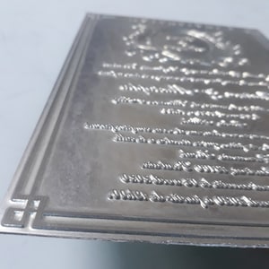 Puede incluir: Un sello de metal plateado con un borde decorativo y letras intrincadas. El sello está en blanco y listo para ser utilizado para crear diseños personalizados.