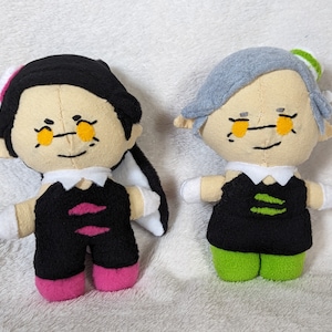 May include: Two plush dolls of characters from a video game. One doll has black hair and a black outfit with pink accents. The other doll has gray hair and a black outfit with green accents. Both dolls have yellow eyes and are on a white surface.
