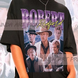 May include: A black t-shirt with a vintage-style graphic print of Robert Redford. The graphic features multiple images of Redford in different poses, with the text "ROBERT REDFORD" in purple letters.