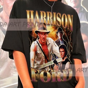 May include: Black t-shirt with a graphic print of Harrison Ford as Indiana Jones. The graphic features a collage of images from the Indiana Jones movies, including the whip, the hat, and the Ark of the Covenant. The text "HARRISON FORD" is printed in gold letters on the shirt.