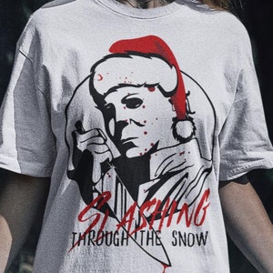 Könnte beinhalten: Weißes T-Shirt mit einem Aufdruck einer Person, die einen Weihnachtsmann-Hut trägt und ein Messer hält. Der Aufdruck lautet "Slashing Through The Snow".