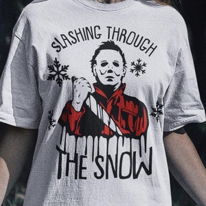 Könnte beinhalten: Weißes T-Shirt mit einem Motiv eines maskierten Mannes, der ein Messer hält, und dem Text "Slashing Through The Snow".