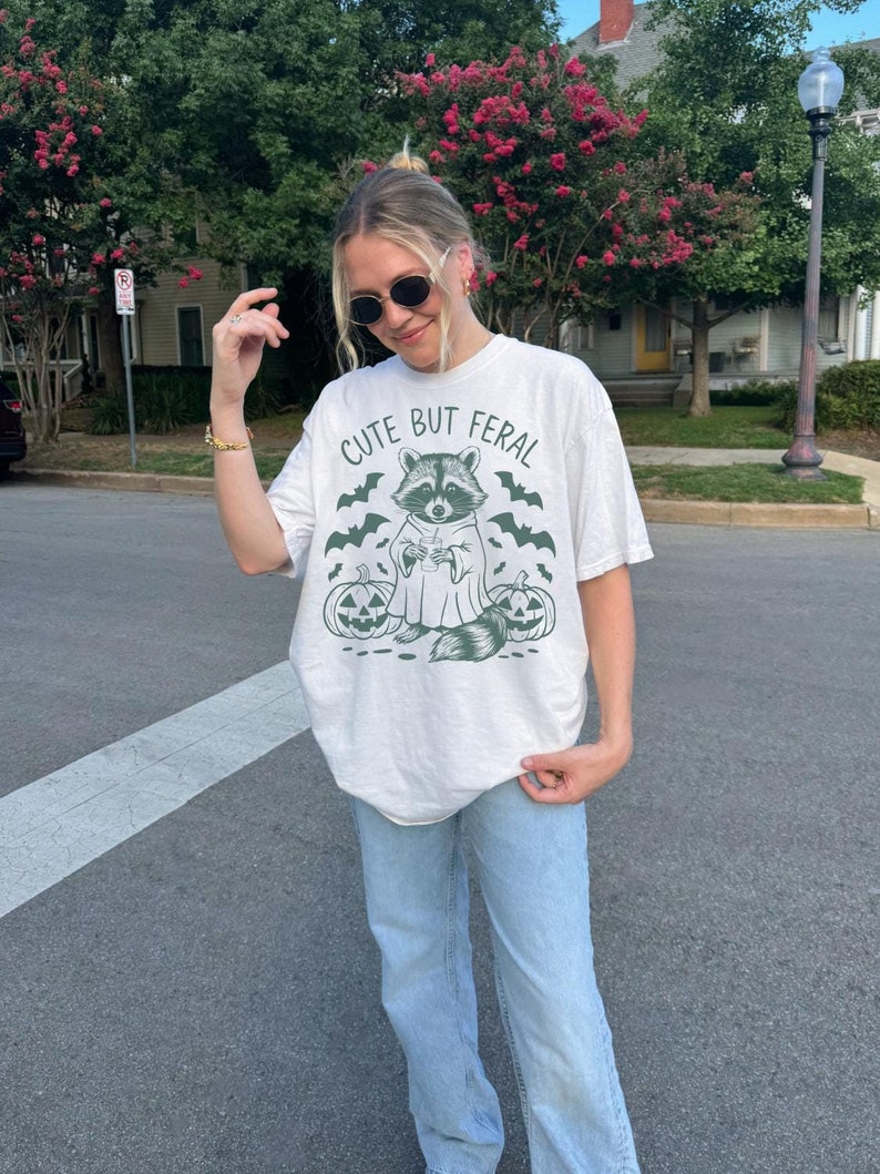 Pu&ograve; includere: Una maglietta bianca con una grafica verde di un procione che indossa un costume da fantasma e tiene una tazza. Il testo "Cute But Feral" &egrave; stampato sopra il procione. La maglietta &egrave; indossata da una persona con jeans blu.