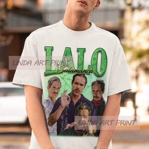May include: White t-shirt with a green and white graphic print of the text "Lalo Salamanca" and four men's faces.