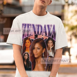 Op de afbeelding: Wit T-shirt met een paarse en witte grafische print van Zendaya. De grafiek bevat meerdere afbeeldingen van Zendaya in verschillende poses. De tekst "Zendaya" is in grote, vetgedrukte letters bovenaan de grafiek gedrukt.