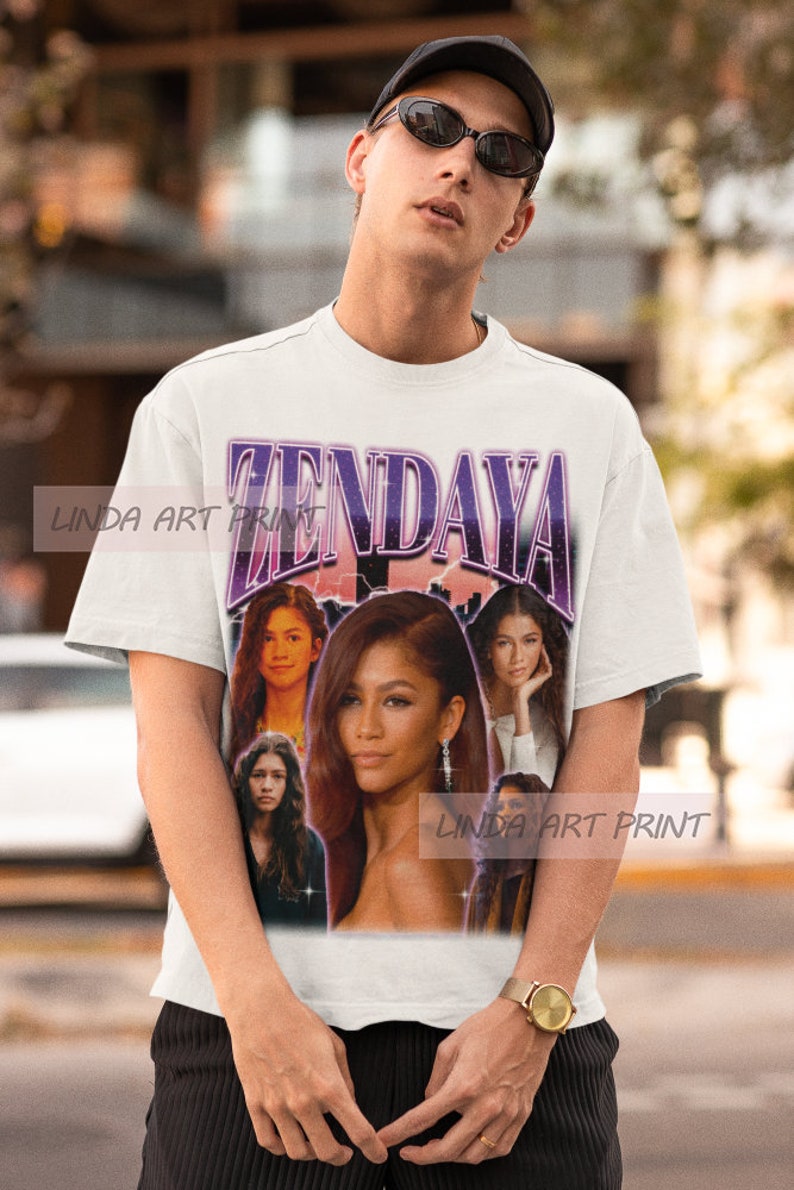 Op de afbeelding: Wit T-shirt met een paarse en witte grafische print van Zendaya. De grafiek bevat meerdere afbeeldingen van Zendaya in verschillende poses. De tekst "Zendaya" is in grote, vetgedrukte letters bovenaan de grafiek gedrukt.