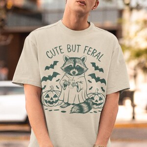 Pu&ograve; includere: Una maglietta beige con una grafica di un procione che indossa un costume da fantasma e tiene un drink. Il testo "Cute But Feral" &egrave; stampato sopra il procione. Due zucche di jack-o'-lantern sono su entrambi i lati del procione.