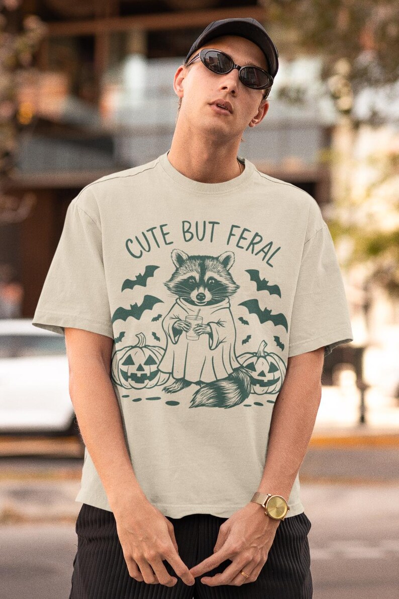 Pu&ograve; includere: Una maglietta beige con una grafica di un procione che indossa un costume da fantasma e tiene un drink. Il testo "Cute But Feral" &egrave; stampato sopra il procione. Due zucche di jack-o'-lantern sono su entrambi i lati del procione.