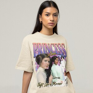 May include: A beige t-shirt with a graphic print of Princess Leia from Star Wars. The graphic features a purple and pink background with the text "Princess Leia" in gold lettering. The graphic also includes a collage of images of Princess Leia in different scenes from the Star Wars movies. The text "Fight Like a Princess" is printed below the graphic.