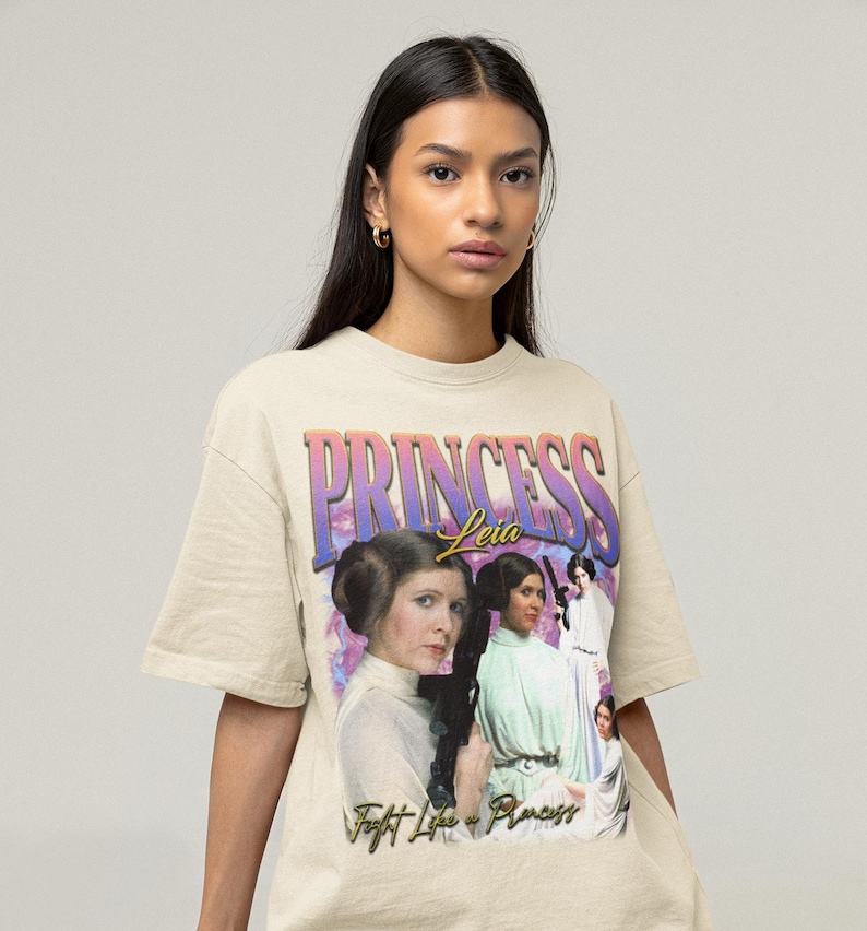 May include: A beige t-shirt with a graphic print of Princess Leia from Star Wars. The graphic features a purple and pink background with the text "Princess Leia" in gold lettering. The graphic also includes a collage of images of Princess Leia in different scenes from the Star Wars movies. The text "Fight Like a Princess" is printed below the graphic.